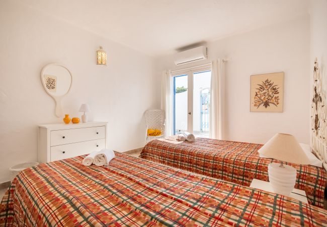 Quarto luminoso com duas camas de solteiro e acesso à varanda Quarto com duas camas de solteiro, cômoda branca, cadeira de vime e portas azuis que dão para o exterior.
