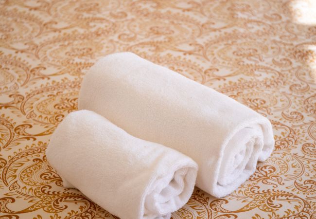 Close-up de toalhas numa cama estampada Uma imagem de close-up de duas toalhas brancas enroladas descansando sobre uma colcha com padrão dourado.