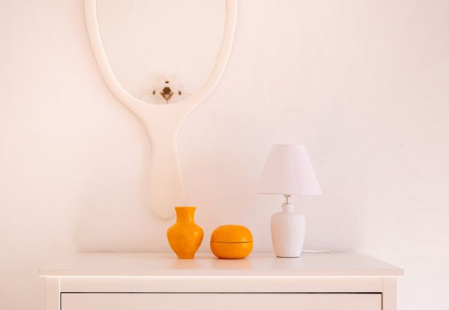 Close-up de uma cômoda com espelho, candeeiro e objetos laranjas Uma cômoda branca com um espelho oval, um candeeiro branco e dois vasos laranjas.