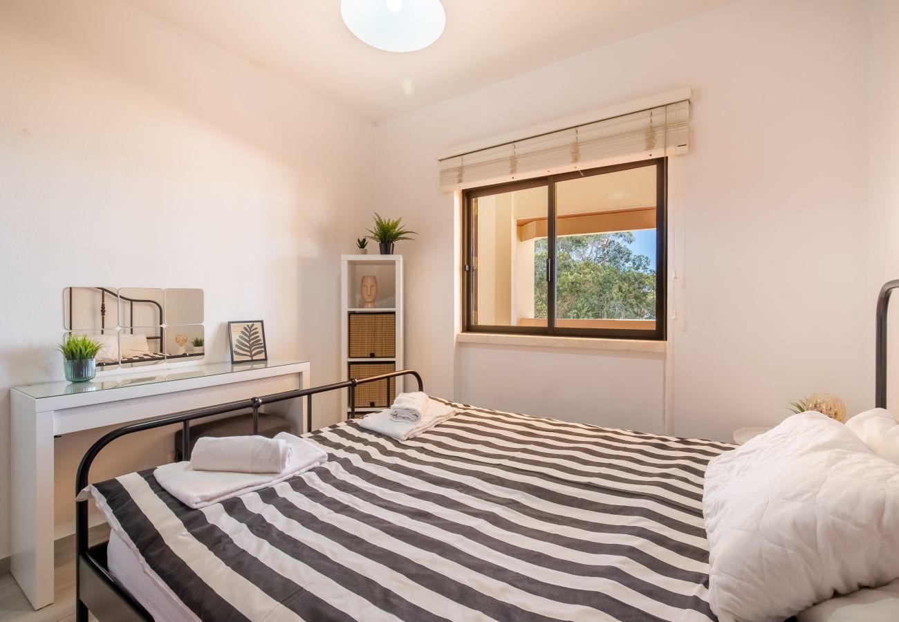 Quarto Claro com Cama Listrada e Vista para a Natureza Interior de quarto luminoso com cama de casal, roupa de cama listrada, secretária branca e janela grande.