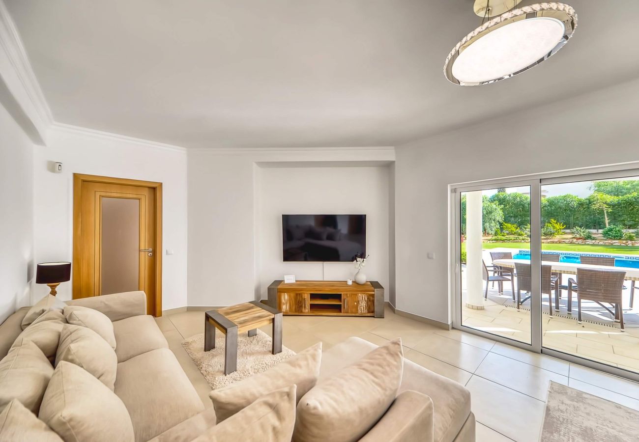 Sala de estar da Villa Larissa com vista para a piscina e o jardim  Sala de estar ampla e luminosa com sofá grande, televisão e porta de vidro para o terraço