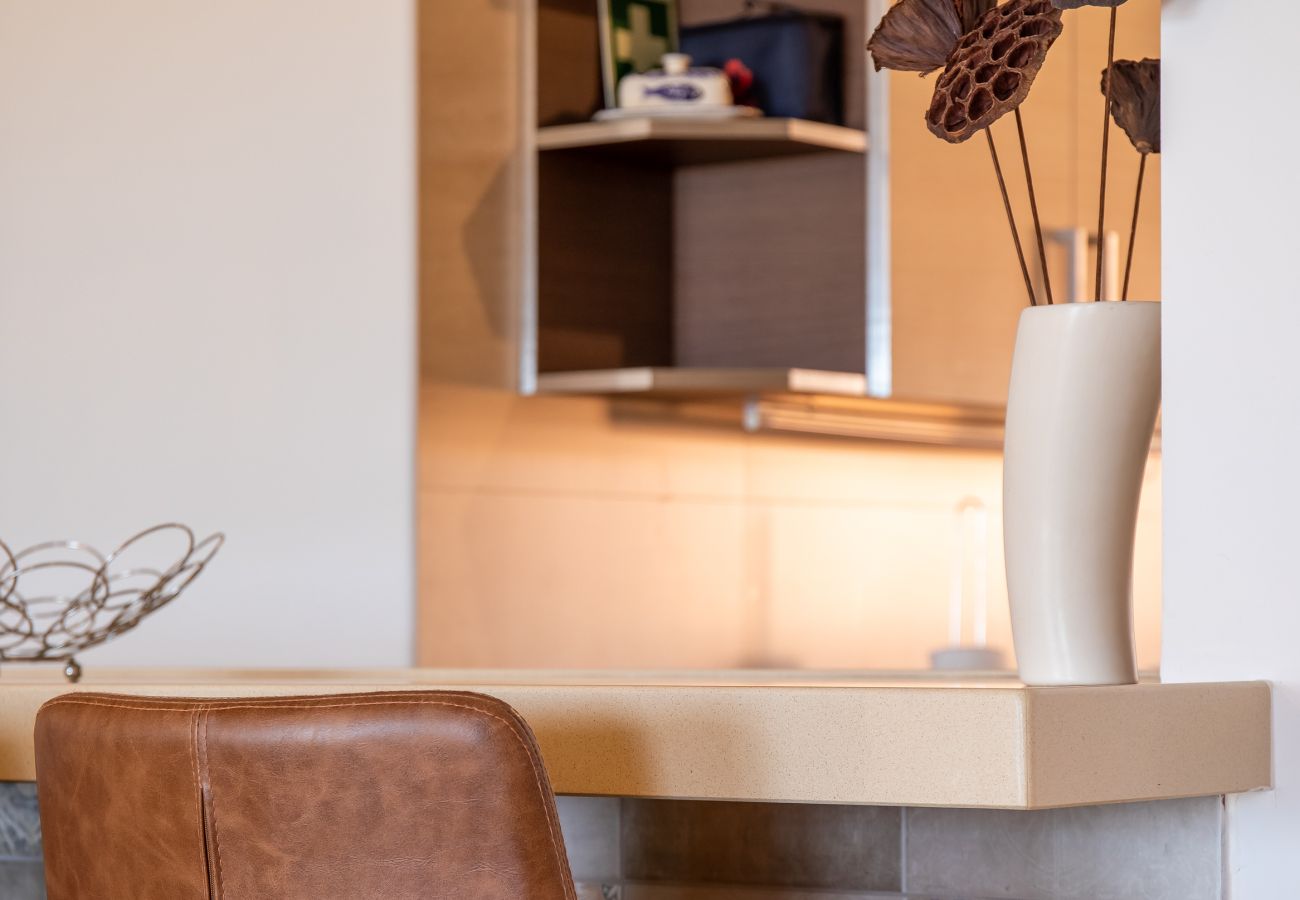 Detalhe Elegante na Área de Estar em Plano Aberto Close-up de um banco de bar de couro castanho, taça decorativa de metal e um vaso com flores secas num balcão de cor clara.