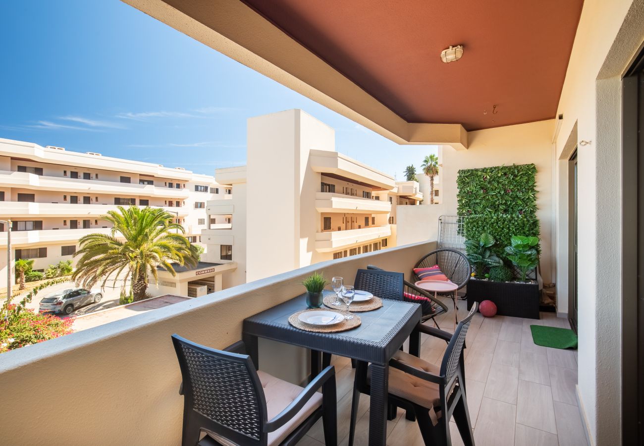 Sonniges Frühstücken auf dem Balkon mit Palmenblick Balkon mit Tisch für vier Personen und Pflanzenwand, mit Blick auf helle Gebäude und eine Palme unter blauem Himmel.